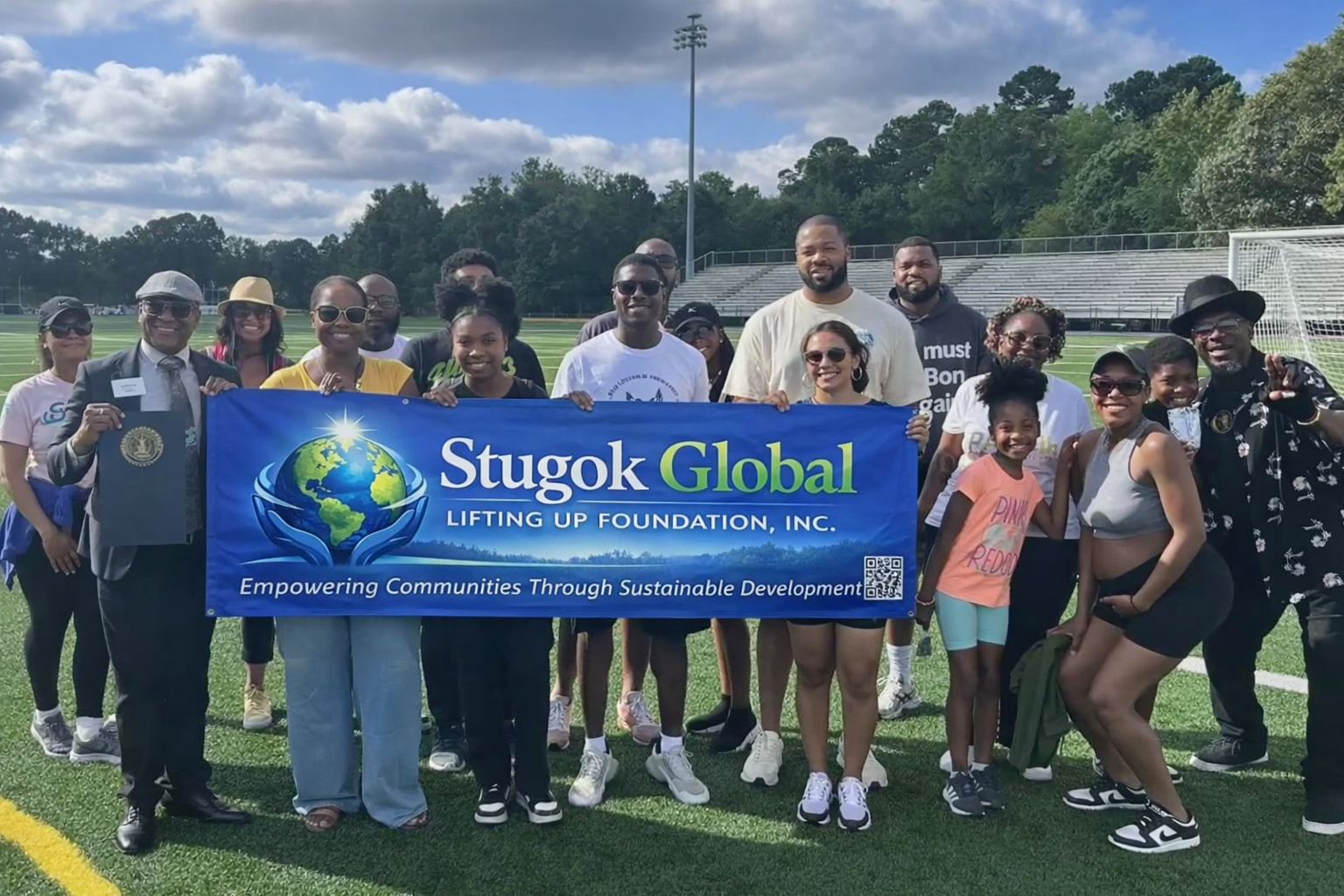Large group of diverse people standing together on a green outdoor sports field holding a Stugok Global banner